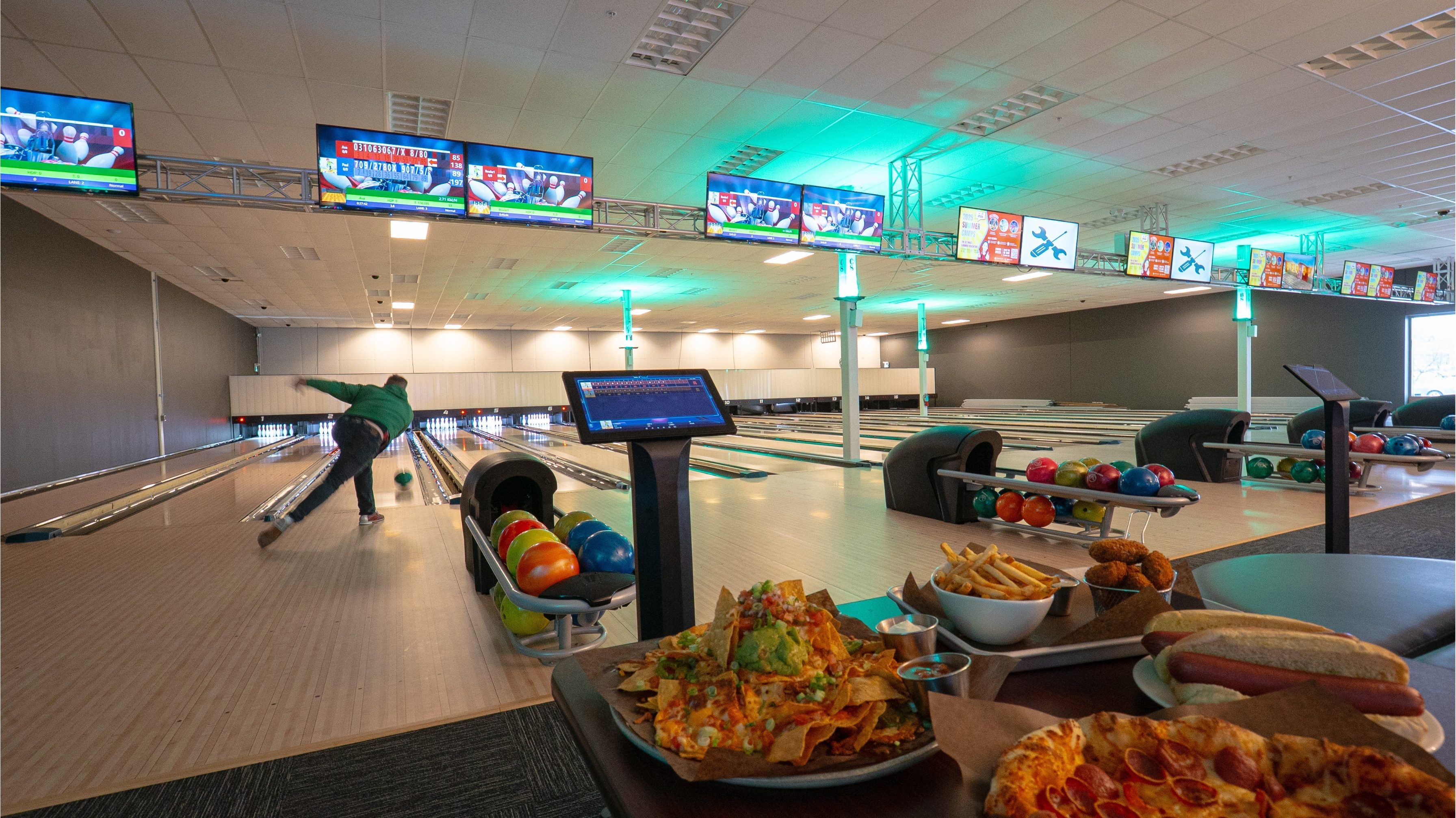 Bowling lanes at Volt Raceway with vibrant lighting, score screens, and a table of food including pizza, nachos, fries, and hot dogs in the foreground.